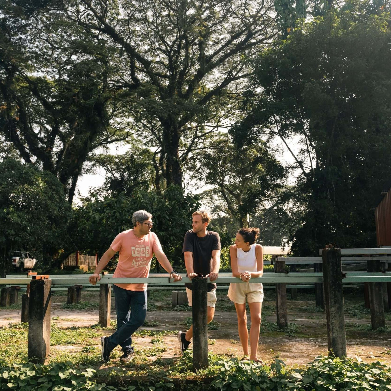 Three people enjoying a sunny day outdoors in a lush green setting, surrounded by trees and rustic benches.