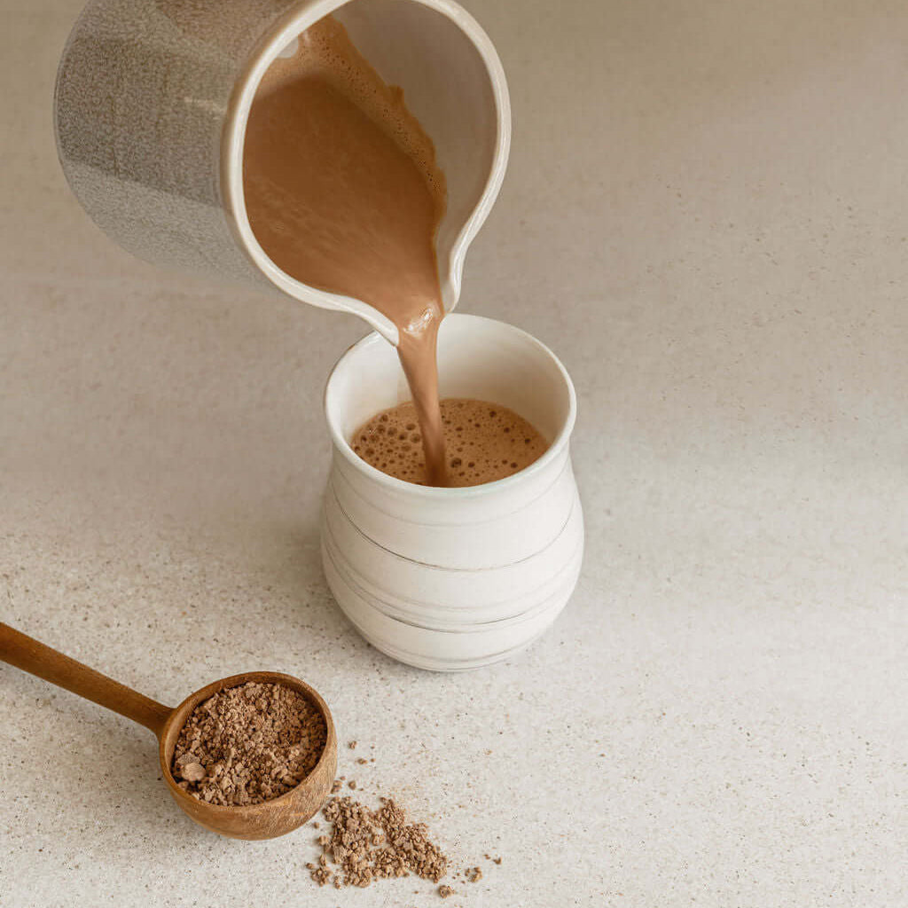 Pouring ceremonial cacao drink into a cup beside cacao powder on a countertop.