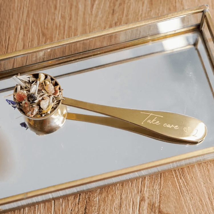 Brass tea scoop engraved with "Take Care," displayed with herbal tea, perfect for ceremonial cacao and premium quality tea brewing.