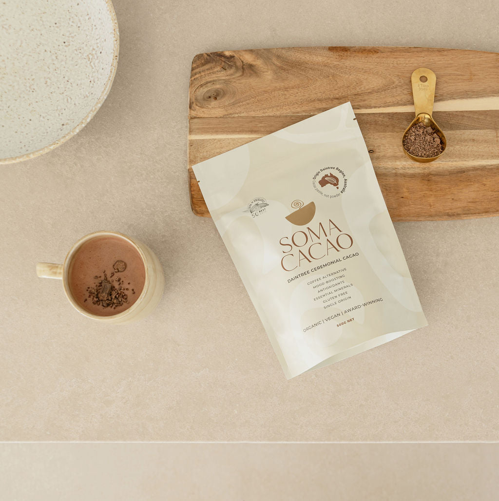 Premium Soma Ceremonial Cacao packaging next to a cup of cacao and a measuring spoon on a wooden board.