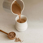 Pouring ceremonial cacao drink into a cup beside cacao powder on a countertop.