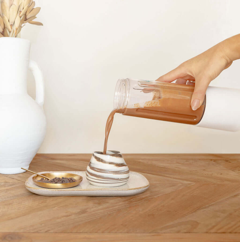 Hand pouring ceremonial cacao from a Soma Portable Blender into a stylish cup on a wooden table.