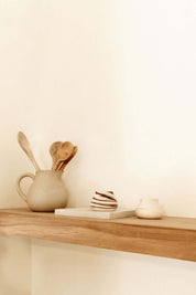 Wooden shelf displaying ceramic jug, wooden utensils, and minimalist decorative items against a light wall.