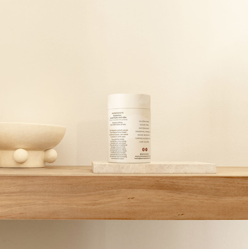 White container with product information on a wooden shelf next to a decorative bowl and stone slab.