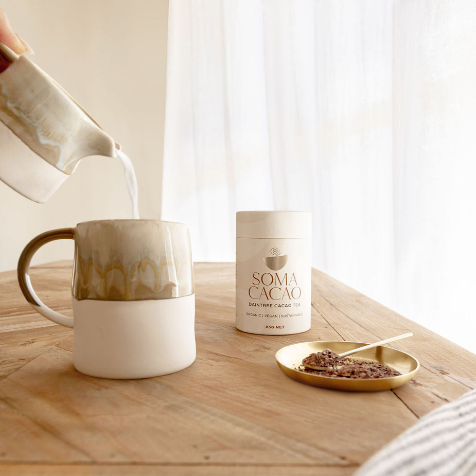 Pouring milk into a mug next to Daintree Cacao Tea canister and a golden dish with cacao sprinkles on a wooden table.