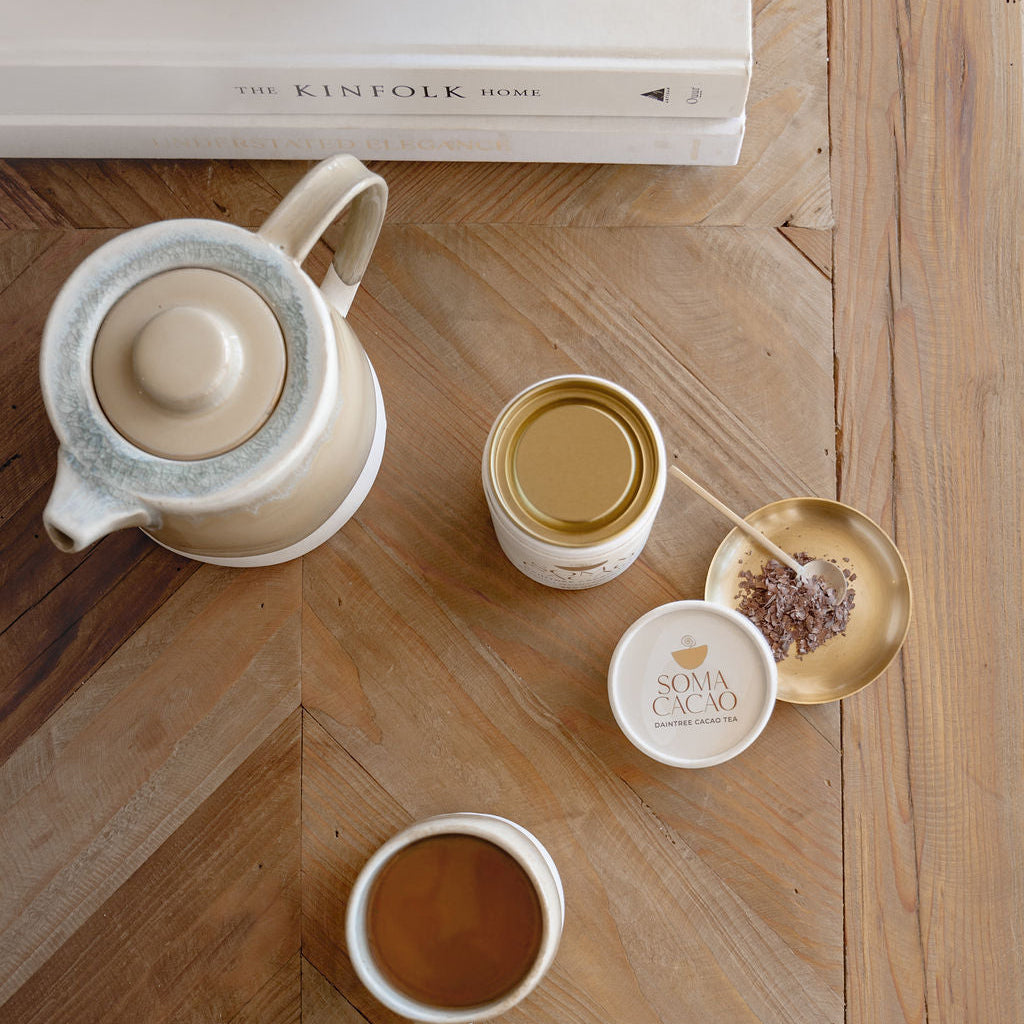 Daintree cacao tea served in a cup with a teapot and premium cacao jar on a wooden table.