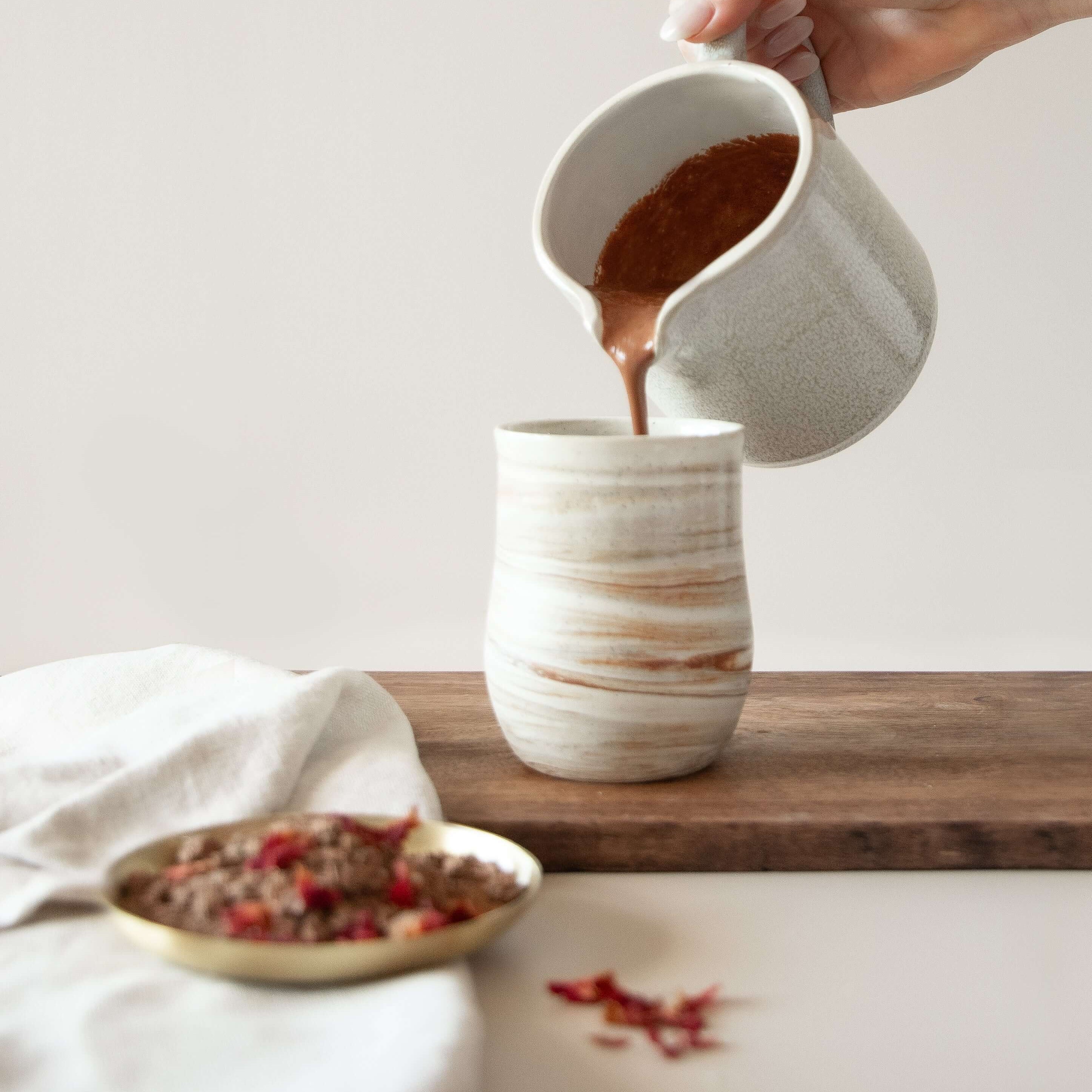 Hand pouring rich ceremonial cacao into a textured cup, showcasing premium quality and warmth in a serene setting.