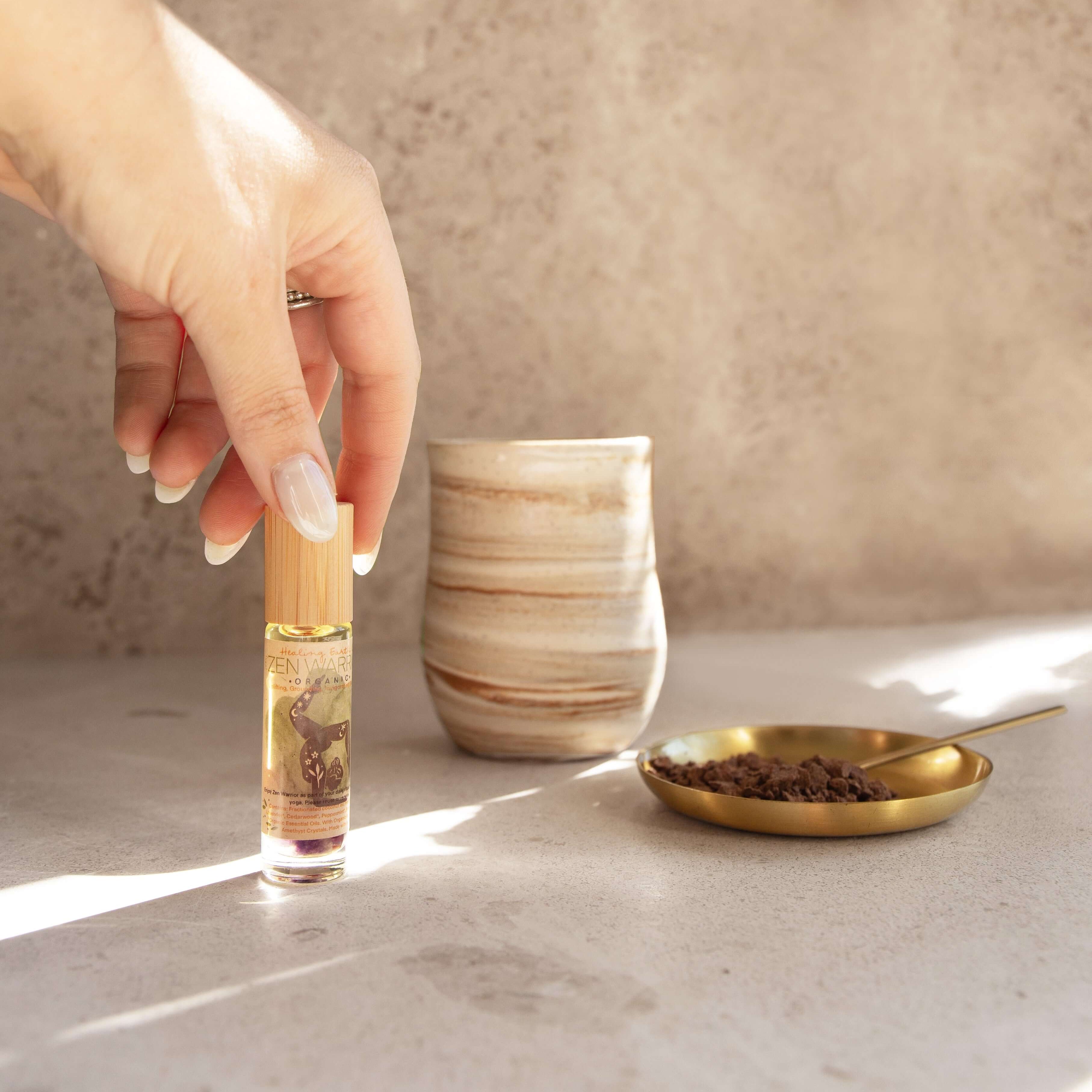 Hand holding Healing Earth Co Zen Warrior perfume roller beside ceremonial cacao and a stylish cup on a serene surface.