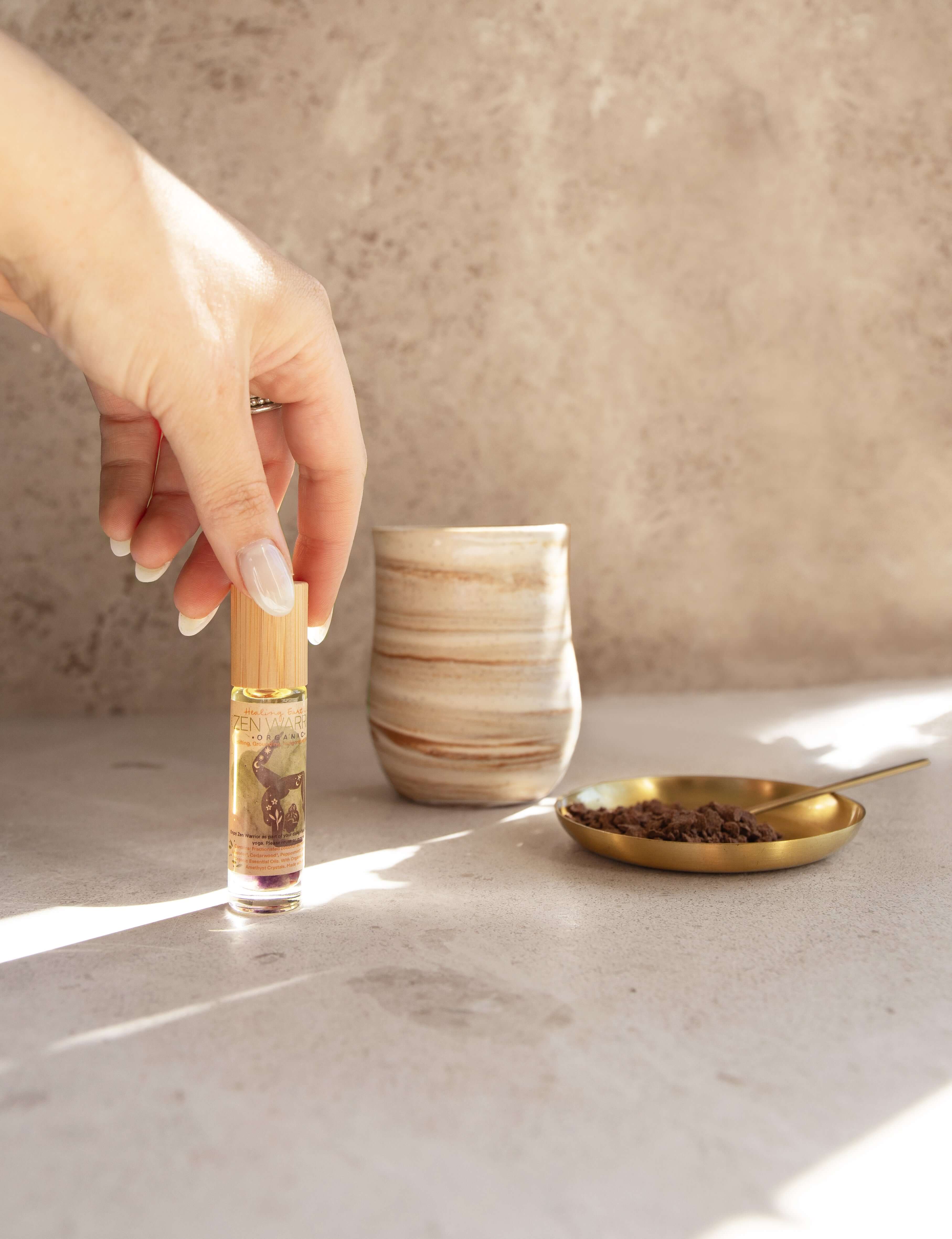 Hand holding Healing Earth Co Zen Warrior perfume roller beside ceremonial cacao and a stylish cup on a serene surface.